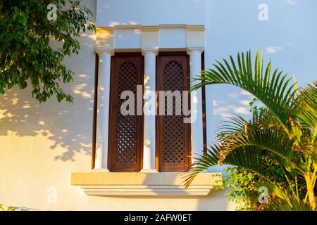 Fenster mit Fensterläden geschlossen. Osten Stil Fenster schließen. Zweigniederlassungen von tropischen Bäumen. Sonnenlicht fällt auf eine Wand mit einem Fenster mit drei Spalten. Stockfoto