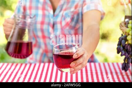 Frau hält Glas Wein oder Traubensaft über Tisch mit roten Tischdecke. In der Nähe befindet sich der Korb der Trauben, in einer Hand eine Kanne. Natürlichen grünen Hintergrund. Stockfoto