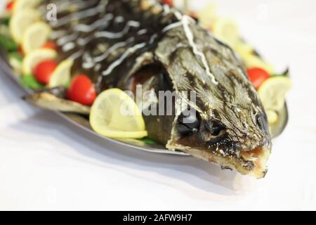 Gebackene Stör mit Tomaten, Salat und Zitronenscheiben. Schwarze Oliven in seinen Augen. Urlaub Teller auf die Platte auf weißer Tisch. Gefüllte Meeresfrüchte. Feier event Party im Restaurant Stockfoto