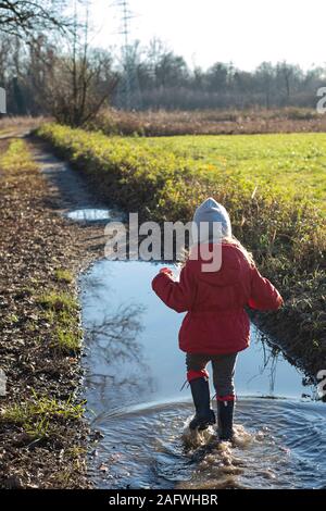 3 Jahre altes Kind Spritzen in eine Pfütze mit roter Jacke und Gummistiefel an einem sonnigen Wintertag. Ansicht von hinten. Vertikale erschossen. Stockfoto