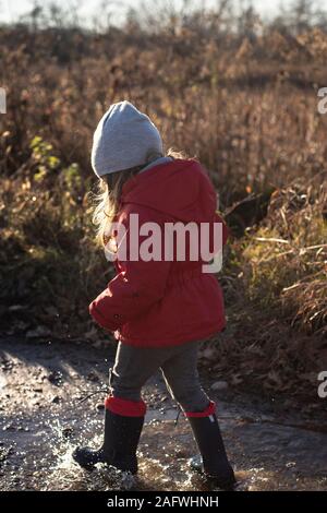 3 Jahre altes Kind Spritzen in eine Pfütze mit roter Jacke und blau Gummi Gummistiefel an einem sonnigen Wintertag. Profil ansehen, vertikal gedreht. Stockfoto