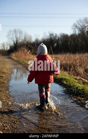 3 Jahre altes Kind Spritzen in eine Pfütze mit Gummistiefel und rote Jacke an einem sonnigen Wintertag. Rückansicht, vertikal gedreht. Stockfoto