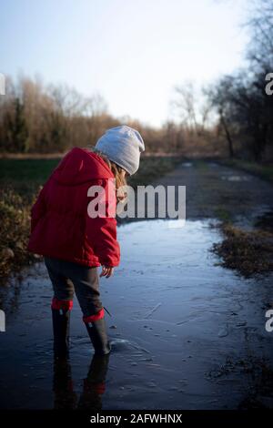 3 Jahre altes Kind Spritzen in eine Pfütze mit roter Jacke und Gummistiefel an einem sonnigen Wintertag. Rückansicht, vertikal gedreht. Stockfoto