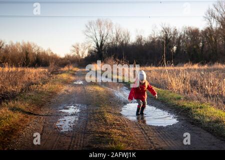 3 Jahre alten Kind in eine Pfütze mit roter Jacke und Gummistiefel springen an einem sonnigen Wintertag. Goldene Stunde Licht. Stockfoto