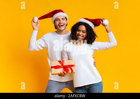 Freudige gemischtrassiges Paar tragen Nikolausmützen und posieren mit Weihnachtsgeschenk Stockfoto