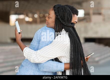 Schwarzer Mann und Mädchen umarmen aber auf Smartphones über die Schulter Stockfoto