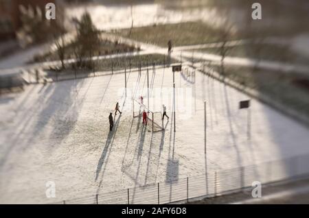 Nicht identifizierte Kinder und ein Erwachsener in der Ferne spielen Fußball in ein schneebedecktes Stadion in der Sonne Tilt Shift Effekt Stockfoto