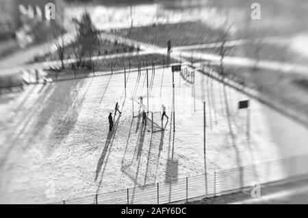 Nicht identifizierte Kinder und ein Erwachsener in der Ferne spielen Fußball in ein schneebedecktes Stadion in der Sonne Tilt Shift Effekt in der schwarz-weiss Stil Stockfoto