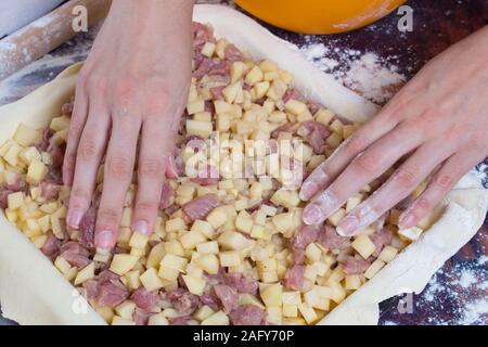 Kochen Kuchen mit Kartoffeln und Fleisch. Frauen Hände legen, schneiden Sie die Kartoffeln und Fleisch auf den gerollten Teig auf dem Tisch liegen. Stockfoto