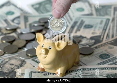 Sparschwein mit zwei Euro Münze in der Hand und in den amerikanischen Dollar usd Banknoten im Hintergrund. Europäische Union Währung und Wirtschaft Konzept Stockfoto