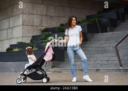 Hübsche, junge Mutter und ihre kleine Tochter in rosa Kinderwagen haben, Spaziergang in der Stadt Stockfoto