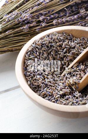 Hölzerne Schüssel von losem, getrocknetem Lavendel Blüten mit Schaufel und Bündel von Stammzellen Blumen im Hintergrund auf weißem Holz beplankt. Stockfoto