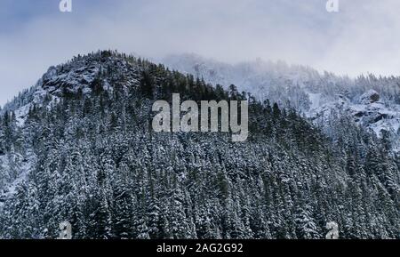 Blauer Himmel kaum sichtbar durch Wolken über einem schneebedeckten, bewaldete Berge. Stockfoto