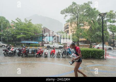 Ao Nang, Krabi/Thailand - Juli 16, 2019: ein Tourist verlangsamende durch starken Regen, der Besitz einer Plastiktüte und ein Paar Flip Flops, entlang der Hauptstraße. Stockfoto