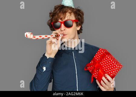 Feier Konzept. Teenager Junge in Geburtstag Kappe und Sonnenbrille isoliert auf Grau mit vorhanden Blasen party Horn lächelnd cool close-up Stockfoto