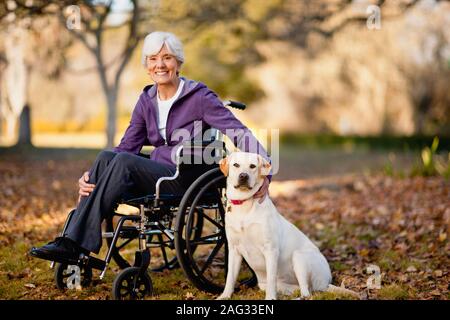 Lächelnde ältere Frau sitzt im Rollstuhl, während Ihr Hund streicheln in einem Park. Stockfoto