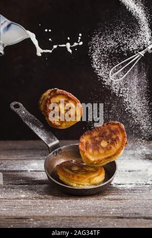 Pfannkuchen und Pfanne schweben über einer Tabelle, und oben ein Schneebesen und bestreut mit Mehl. Milch ist das Gießen von einer Schale Stockfoto