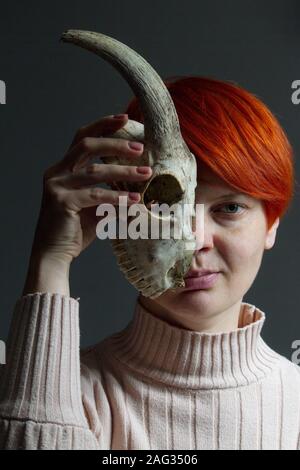 Porträt eines mittleren Alters, rothaarige Frau mit der Hälfte eine Ziege Schädel vor ihrem Gesicht, doppelzüngigkeit Konzept Stockfoto