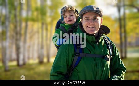 Porträt eines lächelnden Vater seine adorable Baby Sohn in einem Rucksack auf dem Rücken, wie sie Spaß wandern in der kalten Wildnis haben. Stockfoto
