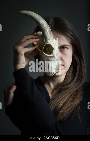 Portrait einer jungen dunkelhaarigen Mädchen Holding eine halbe Ziege Schädel vor ihrem Gesicht, doppelzüngigkeit Konzept Stockfoto