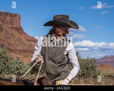Eine junge, attraktive Arbeitsbedingungen cowgirl Wrangler auf Ihrem Pferd auf einer Ranch in der Nähe von Moab, Utah. Hinter ihr sind die sandsteinfelsen der Schlucht der Colora Stockfoto