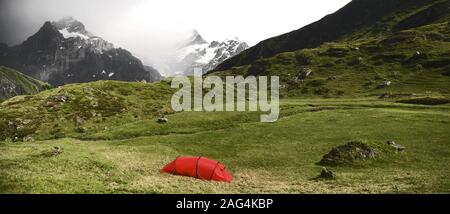 Panoramablick auf ein rotes Zelt auf einem grünen und gelben Gras, hochalpine Berge im Hintergrund Stockfoto