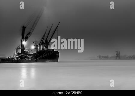 Graustufenaufnahme eines Industrieschiffs mit Licht auf der Meer mit Nebel umhüllt Stockfoto