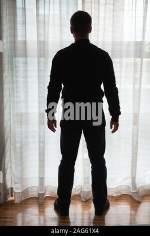 Junge Mann steht in der Nähe von hellen Fenster mit Tüll und sieht außerhalb, Gegenlicht silhouette Foto Stockfoto