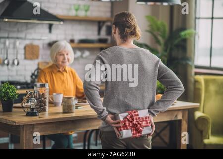 Lassen Sie sich überraschen. Große Mann in Grau shirt versteckt sich ein Geschenk für Mama hinter seinem Rücken Stockfoto