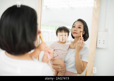 Lustige Familie zu Hause. Mutter und Kind Mädchen tun Sie Ihr Make-up und Spaß haben in der Nähe von Spiegel. Baby Mädchen erforscht die Mutter Kosmetik zu Hause Stockfoto