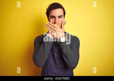 Junge schöne Ladenbesitzer Mann mit Schürze stehend über isolierte gelben Hintergrund schockiert über den Mund mit Händen für Fehler. Geheime Konzept. Stockfoto