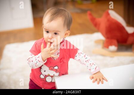 Süße 1 jährige Mädchen beißt Finger, Konzept der Kinderkrankheiten. Mädchen in hello kitty Red Shirt stehend Stockfoto