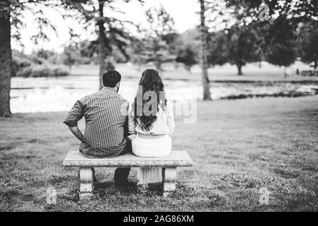 Graustufenaufnahme von hinten eines Paares auf einem sitzend Bank im Park mit einem geröbten Hintergrund Stockfoto