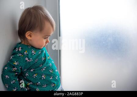 Cute kaukasischen ein Jahr alten Baby Mädchen in grünes T-Shirt mit Rücken an der Wand neben der hellen Fenster. Stockfoto