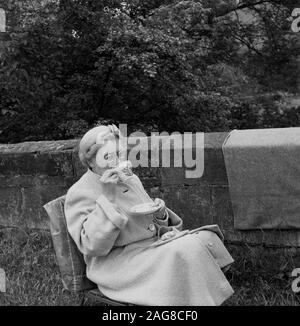 1960er Jahre, historisch, eine reife Dame in Mantel und Hut, die draußen auf einem Stuhl neben einer Steinmauer sitzt, eine Tasse Tee und einen Scone hat, England, Großbritannien. Stockfoto