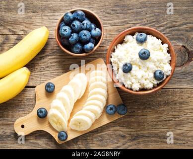 Braun Holz Schale hausgemachter Topfen mit Banane, Marmelade, Blaubeeren auf einem dunklen Braun Holz- Hintergrund, Ansicht von oben, kopieren Raum Stockfoto