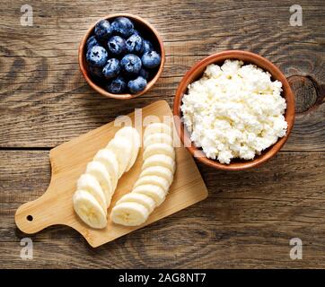 Braun Holz Schale hausgemachter Topfen mit Banane, Marmelade, Blaubeeren auf einem dunklen Braun Holz- Hintergrund, Ansicht von oben, kopieren Raum Stockfoto