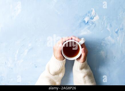 Frau mit Tasse heißen Tee auf einem blauen Tisch, Hände in warmen Pullover mit Becher, Winter morgen Konzept, Ansicht von oben Stockfoto