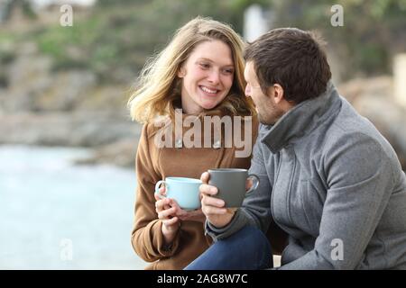 Glückliches Paar von Erwachsenen sprechen trinken Kaffee sitzen im Freien im Winter am Strand Stockfoto