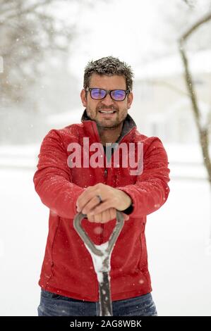 Mann trägt eine rote Winterjacke mit den Händen an Eine Schneeschaufel, während sie zur Kamera lächelt Stockfoto