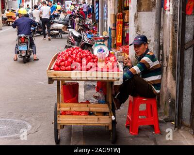 HAIKOU, HAINAN, CHINA - Mar 2 2019 - Street Hersteller raucht eine Zigarette, während Verkauf von tropischen Wachs Äpfel/Jambu Obst von einer Schubkarre Stockfoto