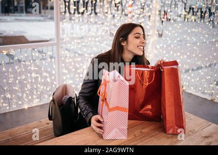 Frau auf der Suche nach Weihnachten Tüten mit Geschenken. Glückliche Frau draußen sitzen am Tisch und Kontrolle Weihnachten Taschen mit Lichtern Mauer umgeben, mit co Stockfoto