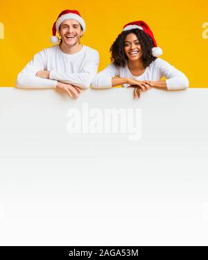 Interracial Paare in Santa Hüte lehnte sich auf weiße Leere Anzeige Brett. Stockfoto