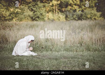 Frau auf dem Boden beim Beten, während sie eine biblische Robe trägt In einem grasbewachsenen Feld Stockfoto