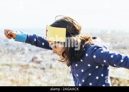 Lustige kleine Kind Mädchen fliegen kraftvoll wie ein Superheld mit der Faust in der Luft getarnt mit hausgemachten Kostüm vor einer Stadtlandschaft, Phantasie Stockfoto