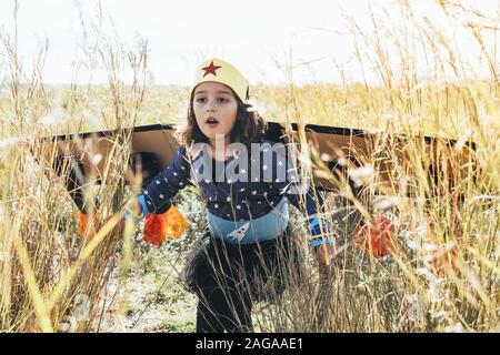 Kind, Mädchen zu Fuß in die Büsche getarnt als ein Superheld mit hausgemachten Kostüm und Karton Flugzeug Flügel, Phantasie und Girl Power Konzept Stockfoto