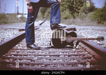 Shallow Focus Aufnahme eines Mannes, der Stiefel trägt und steht Auf Bahngleisen mit einem Fuß auf der Gitarre Stockfoto