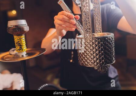 Shisha Schüssel mit Kohle für das Rauchen Nahaufnahme. Hände halten Pinzetten für die Shisha Wasserpfeife und passt die heißen Kohlen in Metall Schüssel. Stockfoto