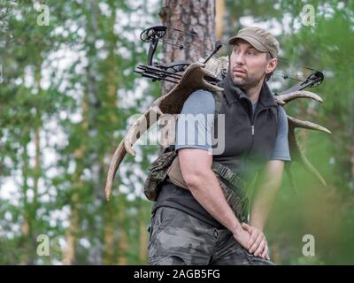 Anzeigen eines Jägers im Sommer Wald mit einem Bogen in den Wald führt Moose Hörner auf seinem Rücken. Stockfoto