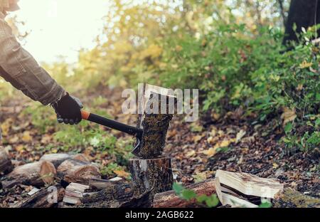 Man schneidet ein Protokoll mit einem industriellen Ax. Teile des Baumes fliegen auseinander. Ein starker Mann hält eine Axt in der Hand auf einem Hintergrund von Wald, Brennholz. Ein Mann lu Stockfoto